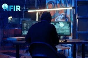 Een persoon met een capuchon op zit aan een bureau met meerdere monitoren waarop code en gegevens worden weergegeven in een schemerig verlichte kamer, met "NFIR Social Engineering" op de muur, waarmee de risico's van phishing-aanvallen in cyberbeveiliging worden benadrukt.